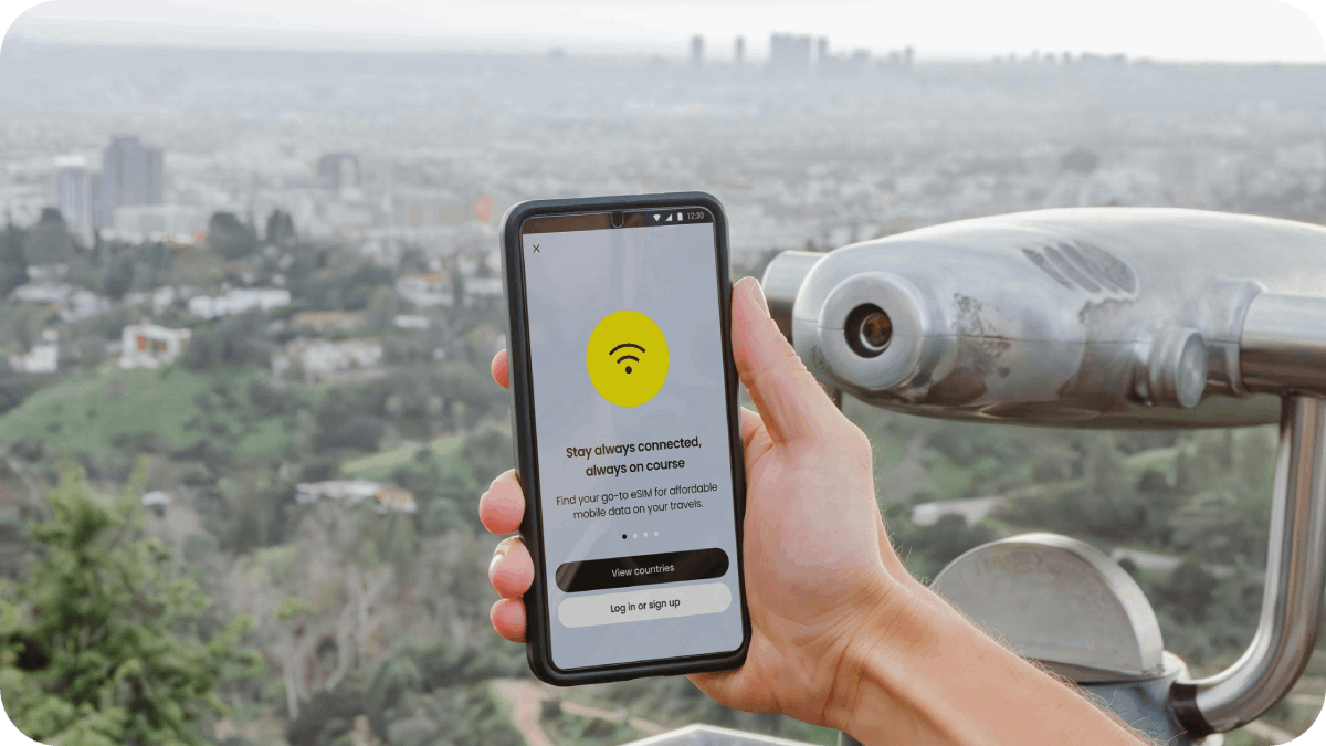 A person holding a smartphone with a connectivity app open, overlooking a cityscape through a public telescope.